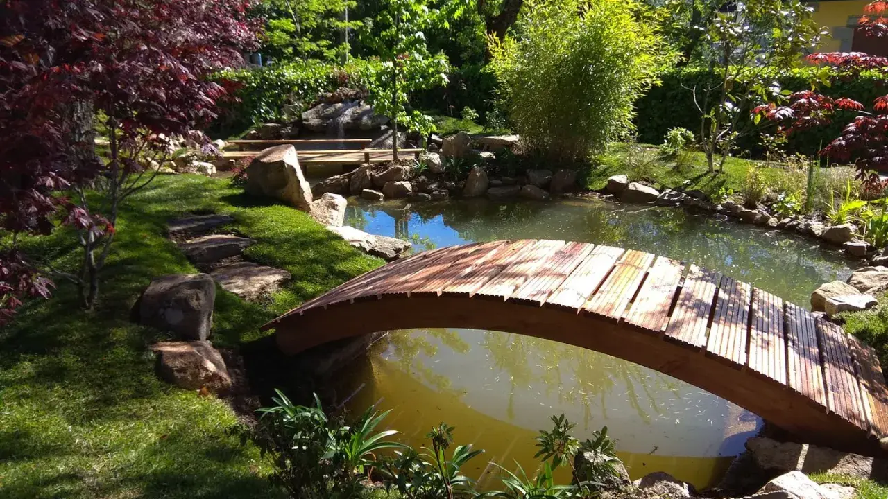 Un puente de madera cruza un estanque, parte de un jardín japonés. Pasos de piedra invitan a explorar.