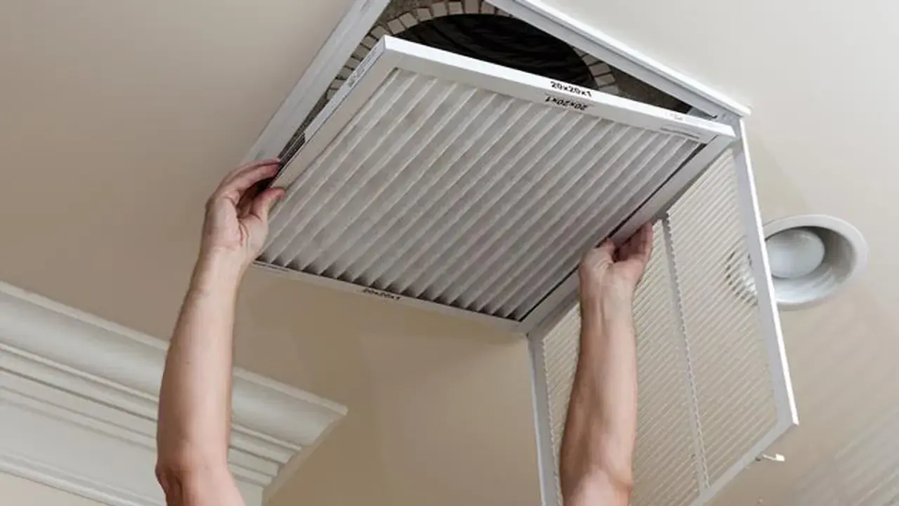 Man hands removing a pleated air filter from a ceiling vent. Essential for limpiar filtros aire acondicionado.