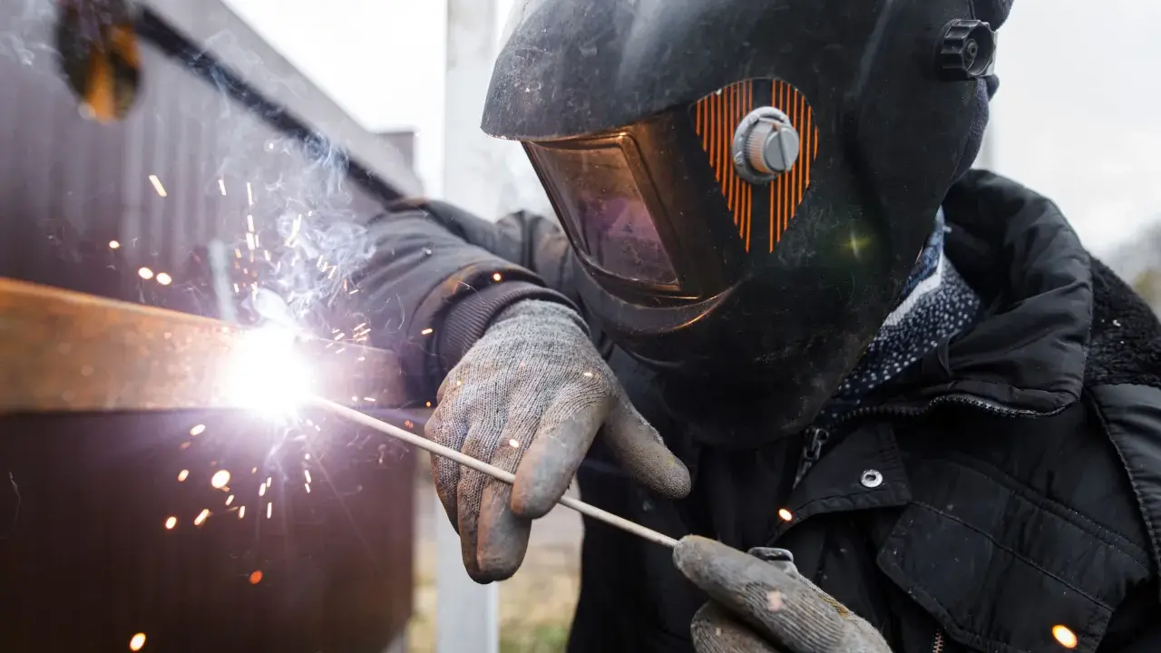 Soldador con casco y guantes usa un electrodo para unir metales, creando chispas brillantes y humo.