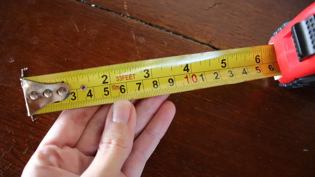 Una mano sujeta una cinta métrica amarilla sobre una mesa de madera. La cinta muestra medidas en pies y metros, demostrando para que sirve la cinta métrica: medir distancias.
