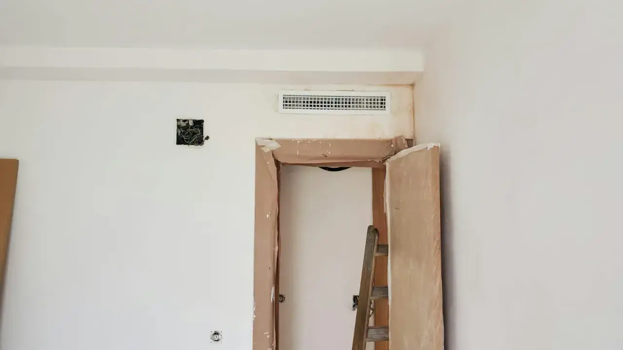Habitación en obra con rejilla de preinstalación aire acondicionado en la pared, puerta abierta y escalera.