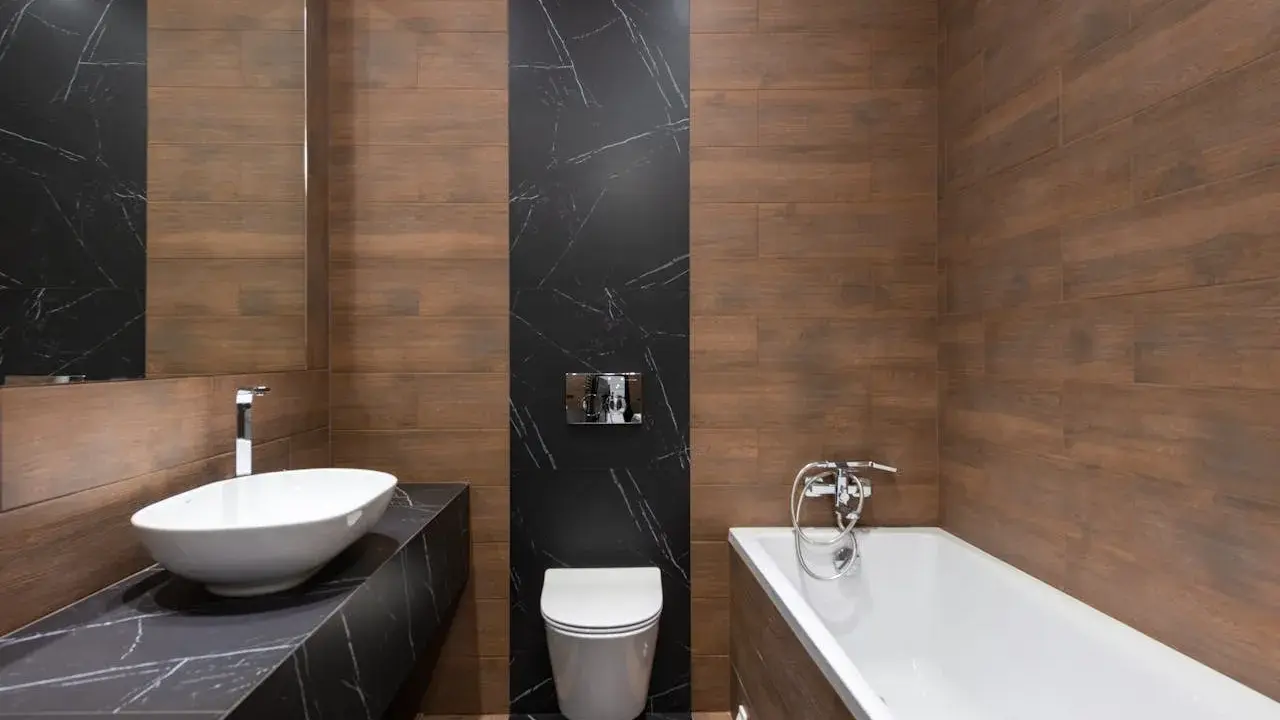 Baño moderno con lavabo ovalado, bañera y un inodoro con salida vertical o horizontal, todo rodeado de azulejos de madera y mármol negro.