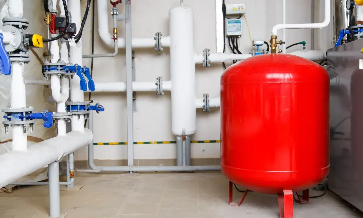 Un sistema de tuberías con válvulas azules y un gran tanque rojo, que es un vaso de expansión, en una sala técnica.