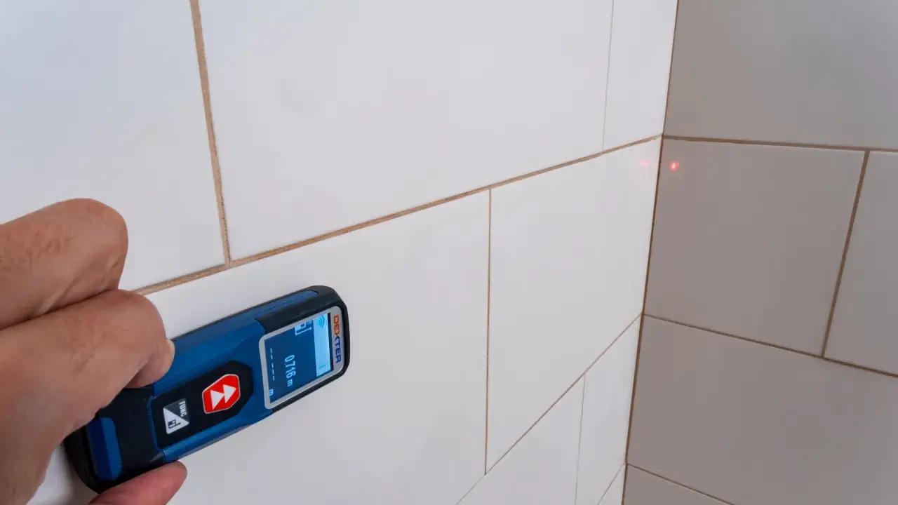 Mano sujetando un aparato para medir con láser rojo apuntando a la esquina de una pared con baldosas blancas.