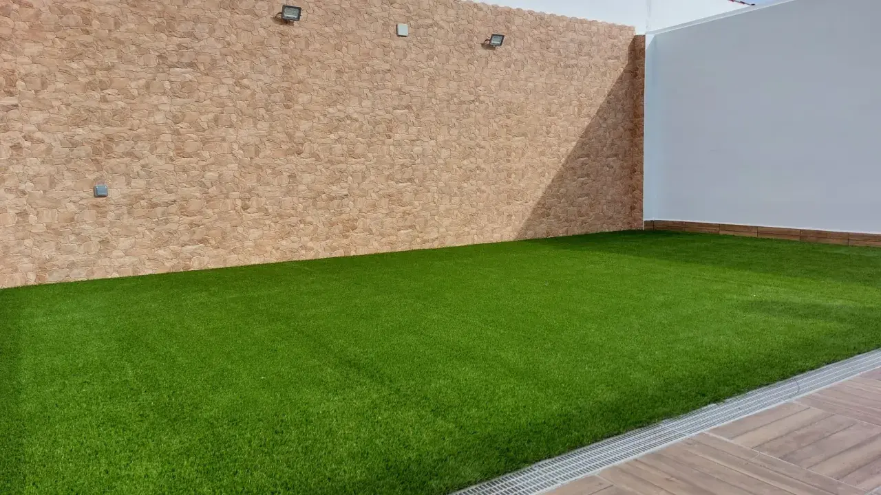 Un patio con césped artificial verde vibrante, paredes de piedra y madera. Ideal para aprender como colocar césped artificial.