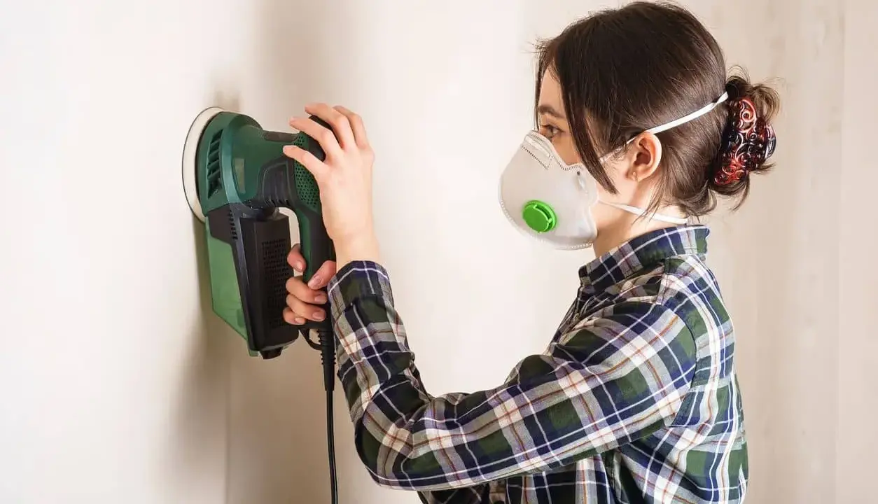 Mujer con mascarilla lijando una pared con lijadora eléctrica.