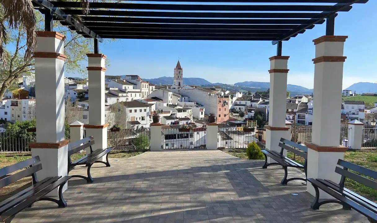 Un mirador con bancos y un arriate de flores ofrece vistas de un pueblo blanco con una torre de iglesia.