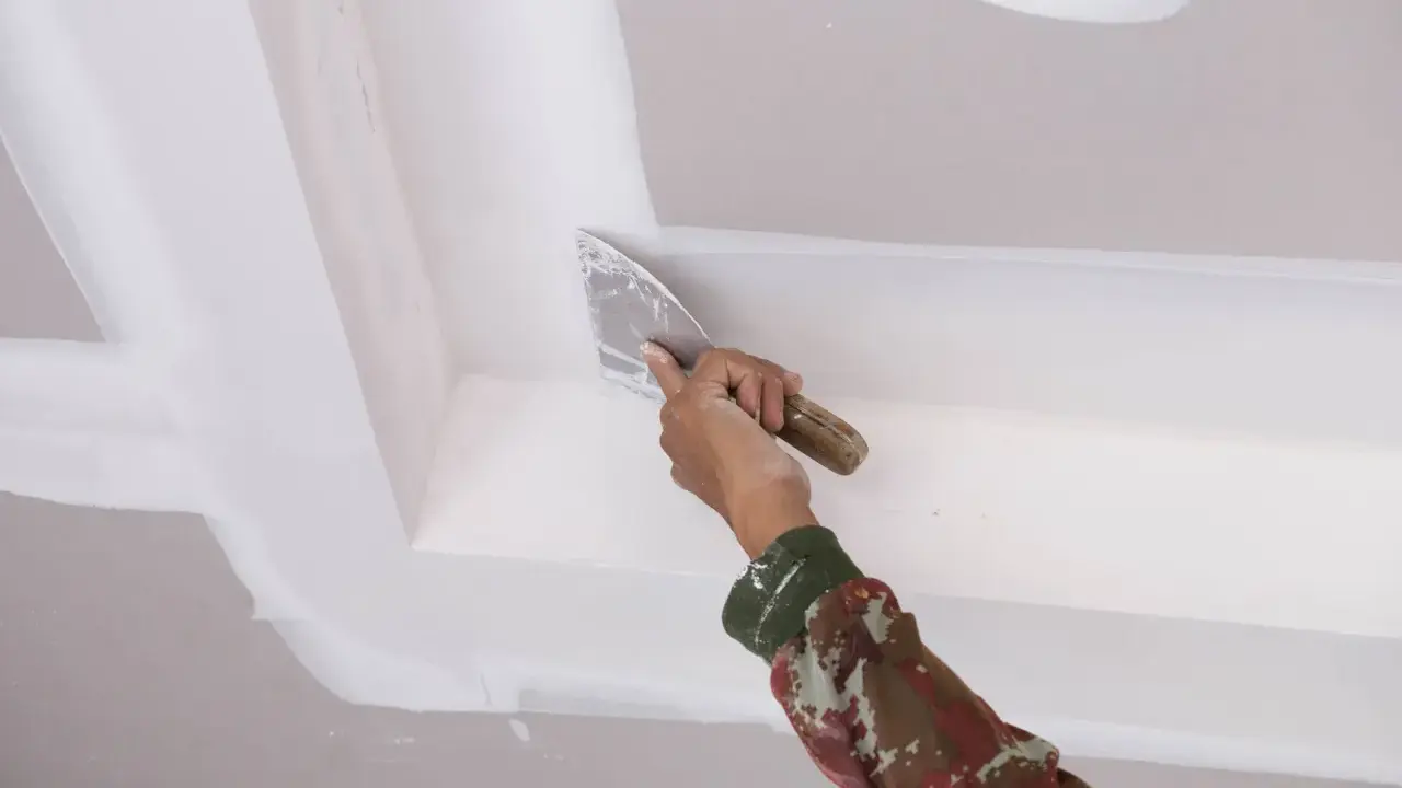 Mano con espátula aplica masilla para reparar techo escayola, dejando un acabado liso y perfecto.