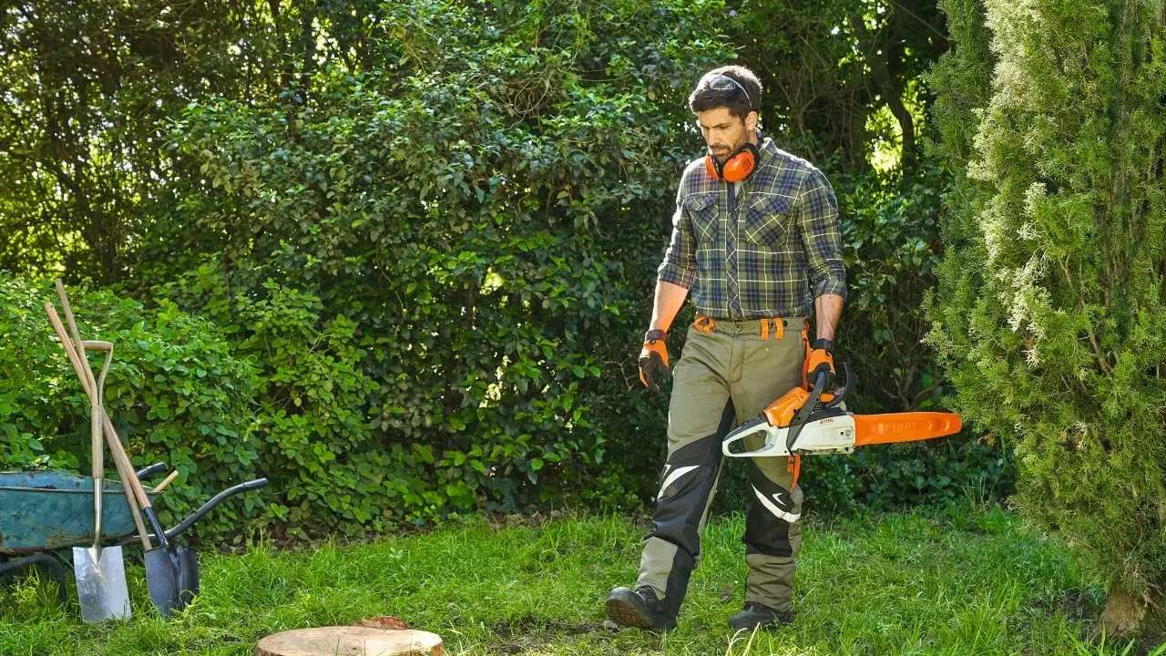 Hombre con motosierra se acerca a un tocon de arbol en un jardín. Herramientas y carretilla a la izquierda.
