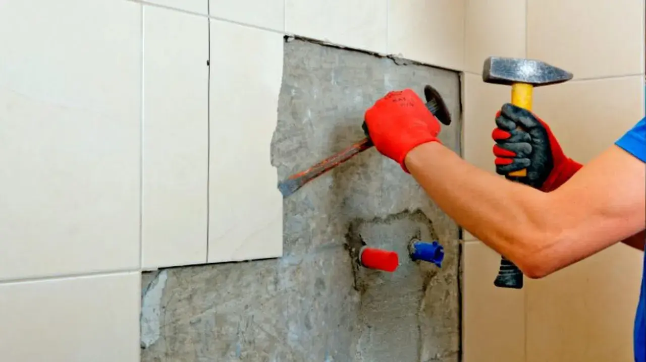 Man con guantes rojos usa un cincel y martillo para quitar azulejos y cemento de una pared.