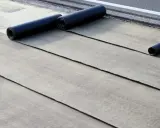 Instalación de tela asfáltica en rollo sobre una superficie. Se aprecian varias capas desplegadas y rollos listos para continuar.