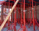 Construcción con varios tipos de puntales rojos sosteniendo una estructura. Un trabajador supervisa.
