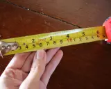 Una mano sujeta una cinta métrica amarilla sobre una mesa de madera. La cinta muestra medidas en pies y metros, demostrando para que sirve la cinta métrica: medir distancias.