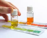 Mano sosteniendo un vial con líquido amarillo sobre tiras de prueba de color para saber la dureza del agua de mi zona. Otro vial con líquido naranja al lado.