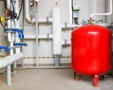 Un sistema de tuberías con válvulas azules y un gran tanque rojo, que es un vaso de expansión, en una sala técnica.