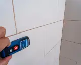 Mano sujetando un aparato para medir con láser rojo apuntando a la esquina de una pared con baldosas blancas.