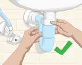 Manos ajustando el sifón azul del lavabo. Un símbolo de verificación verde indica que la tarea de cambiar sifón lavabo está completa.