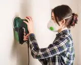 Mujer con mascarilla lijando una pared con lijadora eléctrica.