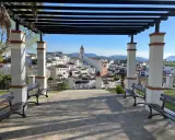 Un mirador con bancos y un arriate de flores ofrece vistas de un pueblo blanco con una torre de iglesia.