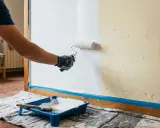 Persona aplicando pintura blanca con rodillo en pared. Se muestra bandeja con pintura y brocha. Aprende como hacer gotele con rodillo.