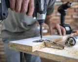 Un hombre usa uno de los tipos de taladros para atornillar una bisagra a un tablero de madera. Otro taladro y herramientas están en la mesa.