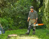 Hombre con motosierra se acerca a un tocon de arbol en un jardín. Herramientas y carretilla a la izquierda.