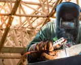 Soldador con máscara y guantes realiza soldadura con electrodo, creando chispas y humo bajo una estructura metálica iluminada por el sol.
