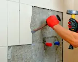 Man con guantes rojos usa un cincel y martillo para quitar azulejos y cemento de una pared.