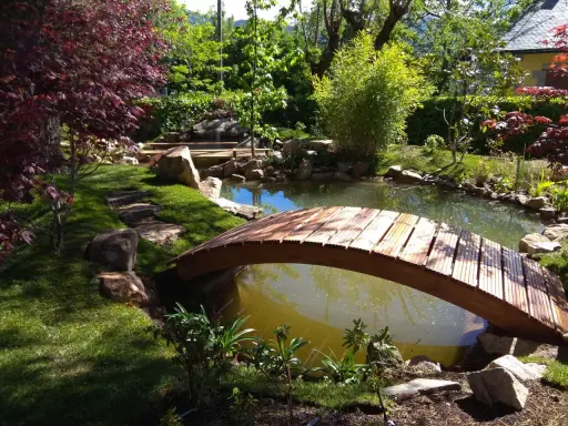 Un puente de madera cruza un estanque, parte de un jardín japonés. Pasos de piedra invitan a explorar.