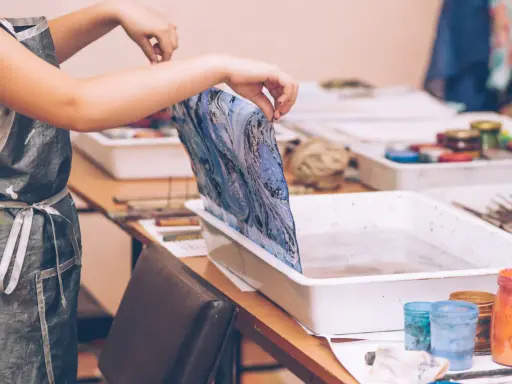 Manos sostienen tela con diseño marmoleado sobre una bandeja con agua, lista para la pintura para mármol.