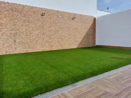 Un patio con césped artificial verde vibrante, paredes de piedra y madera. Ideal para aprender como colocar césped artificial.