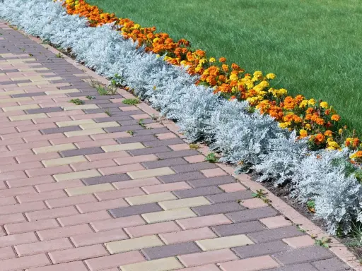 Un camino de adoquines de colores bordea unos parterres de flores anaranjadas y amarillas, con plantas plateadas y césped verde.