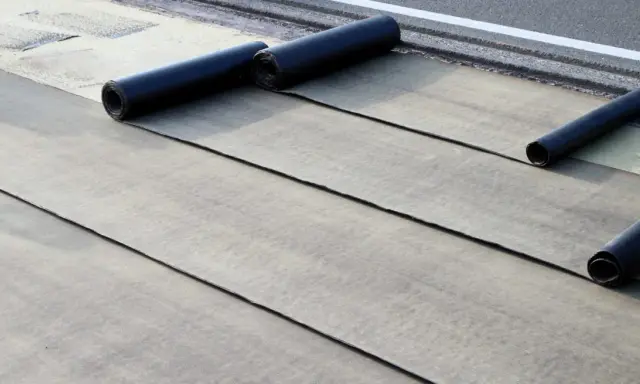 Instalación de tela asfáltica en rollo sobre una superficie. Se aprecian varias capas desplegadas y rollos listos para continuar.