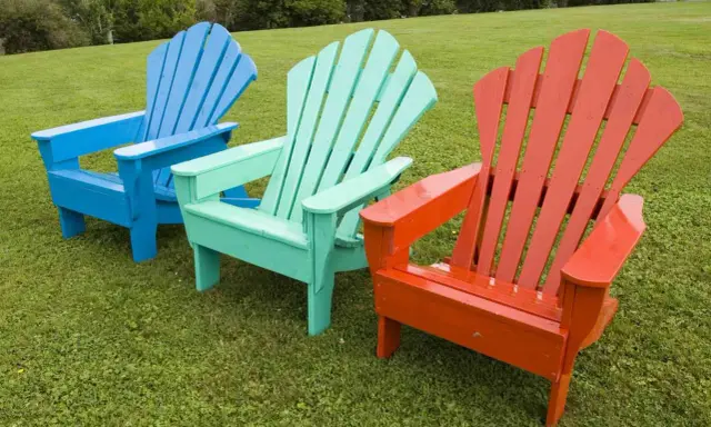 Tres sillas de madera de estilo Adirondack, pintadas de azul, verde menta y naranja, lucen vibrantes sobre el césped. ¡Un proyecto de pintar sillas de madera sin lijar que da vida al jardín!