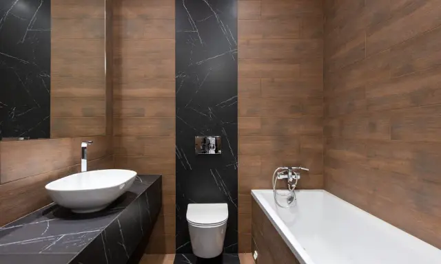 Baño moderno con lavabo ovalado, bañera y un inodoro con salida vertical o horizontal, todo rodeado de azulejos de madera y mármol negro.