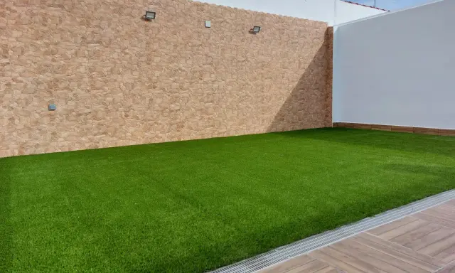 Un patio con césped artificial verde vibrante, paredes de piedra y madera. Ideal para aprender como colocar césped artificial.