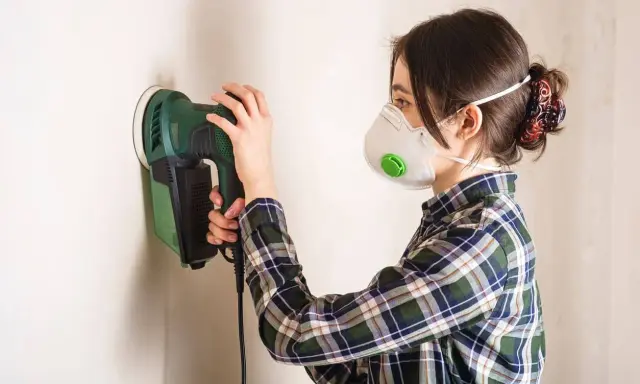 Mujer con mascarilla lijando una pared con lijadora eléctrica.