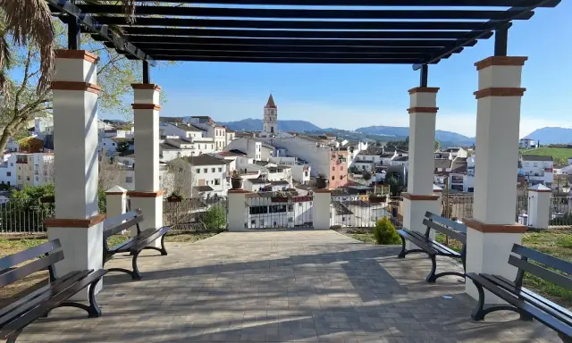 Un mirador con bancos y un arriate de flores ofrece vistas de un pueblo blanco con una torre de iglesia.