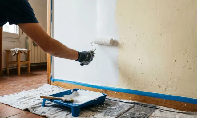 Persona aplicando pintura blanca con rodillo en pared. Se muestra bandeja con pintura y brocha. Aprende como hacer gotele con rodillo.