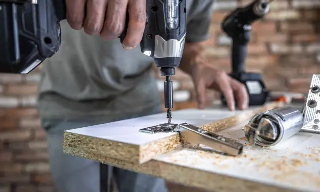 Un hombre usa uno de los tipos de taladros para atornillar una bisagra a un tablero de madera. Otro taladro y herramientas están en la mesa.