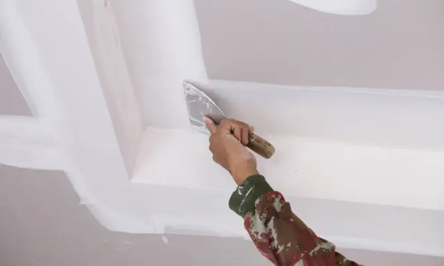 Mano con espátula aplica masilla para reparar techo escayola, dejando un acabado liso y perfecto.