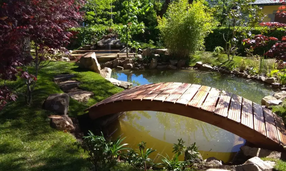 Un puente de madera cruza un estanque, parte de un jardín japonés. Pasos de piedra invitan a explorar.