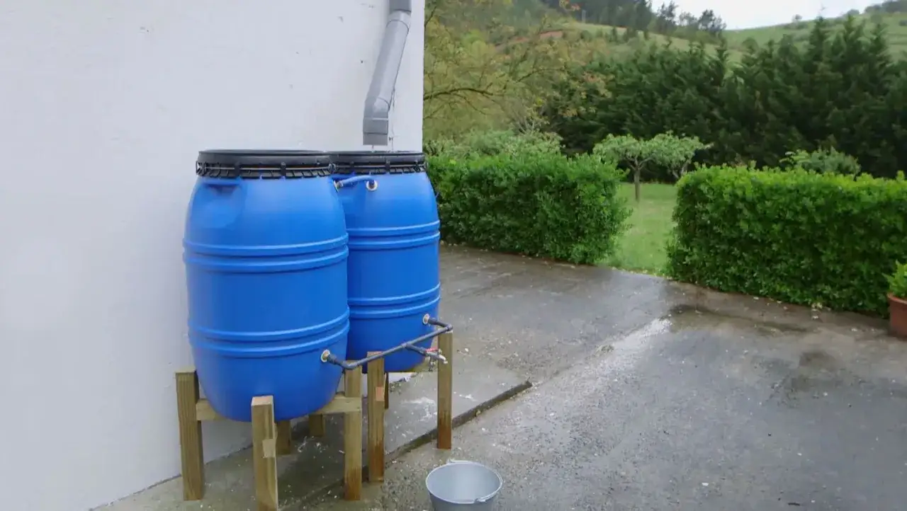 Dos barriles azules forman un dep&oacute;sito de agua de lluvia casero, conectados a un bajante. Un grifo permite el acceso al agua recolectada.