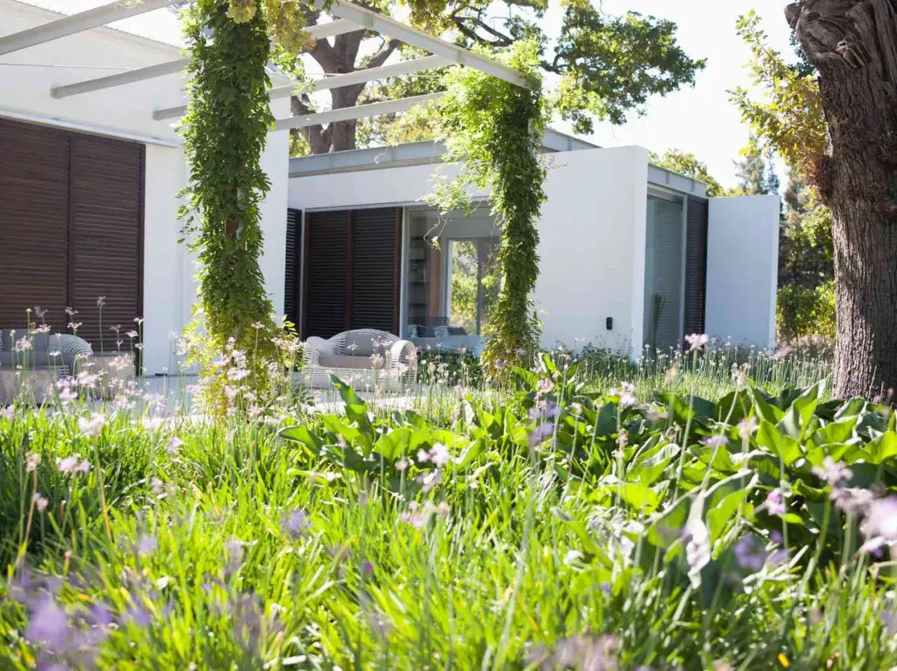 Jardín moderno con exuberante vegetación y flores, ideal para la instalación de césped. Muebles de exterior bajo pérgola.