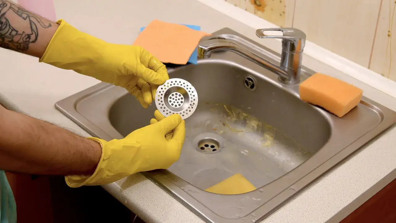 Manos con guantes amarillos sostienen un colador para el esquema del desague de la cocina. Hay una esponja naranja y pa&ntilde;os cerca.