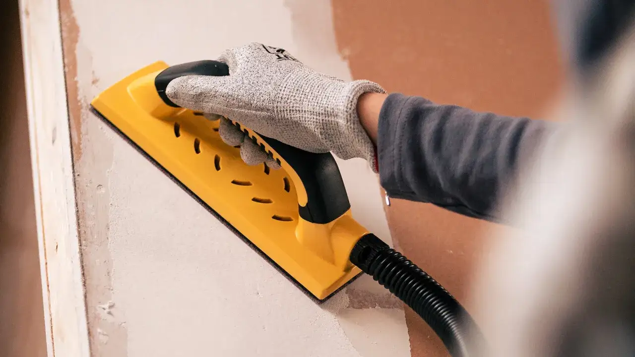 Mano enguantada usando lijadora el&eacute;ctrica amarilla para alisar una pared.