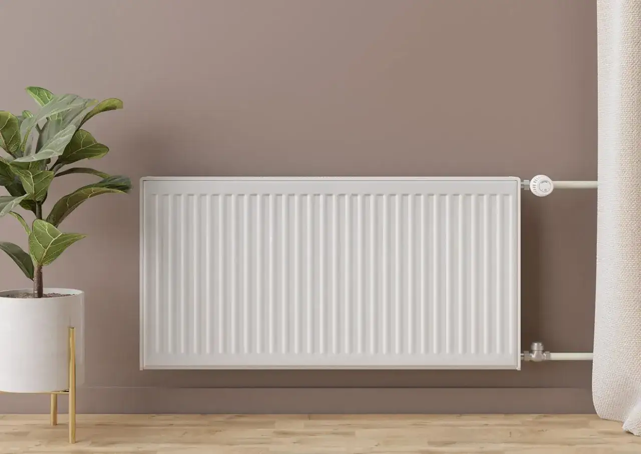 Instalación de radiadores: un radiador blanco moderno junto a una planta en maceta y una cortina, listo para calentar el hogar.