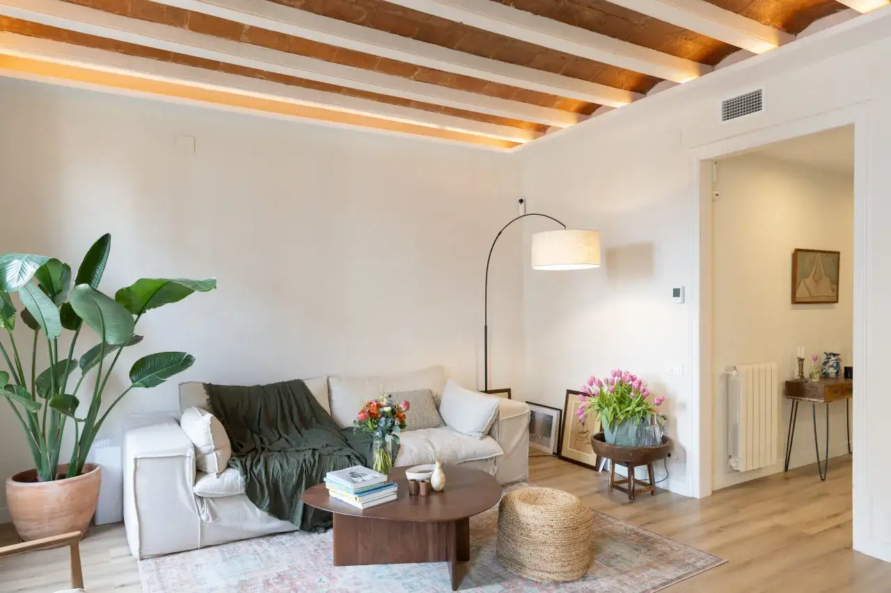 Sal&oacute;n acogedor tras reforma piso peque&ntilde;o. Sof&aacute; beige, manta verde, mesa de centro de madera, plantas y flores. Vigas de madera en el techo.