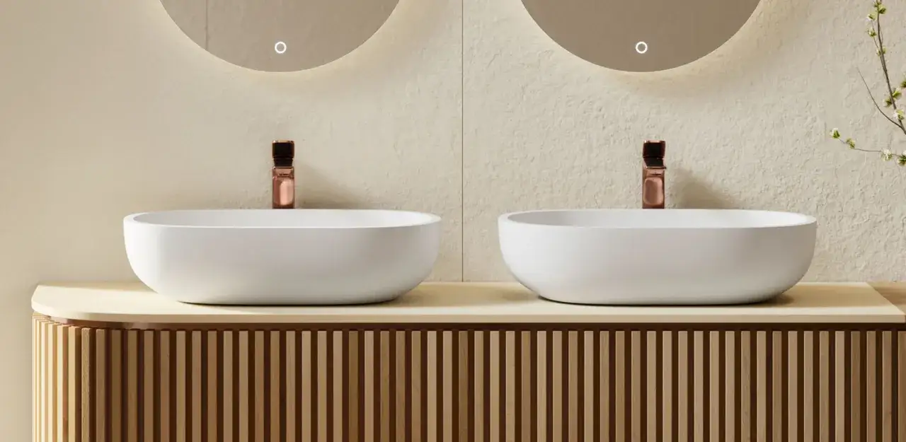 Dos lavabos blancos sobre encimera con grifer&iacute;a cobre. Mueble de ba&ntilde;o con lamas de madera.
