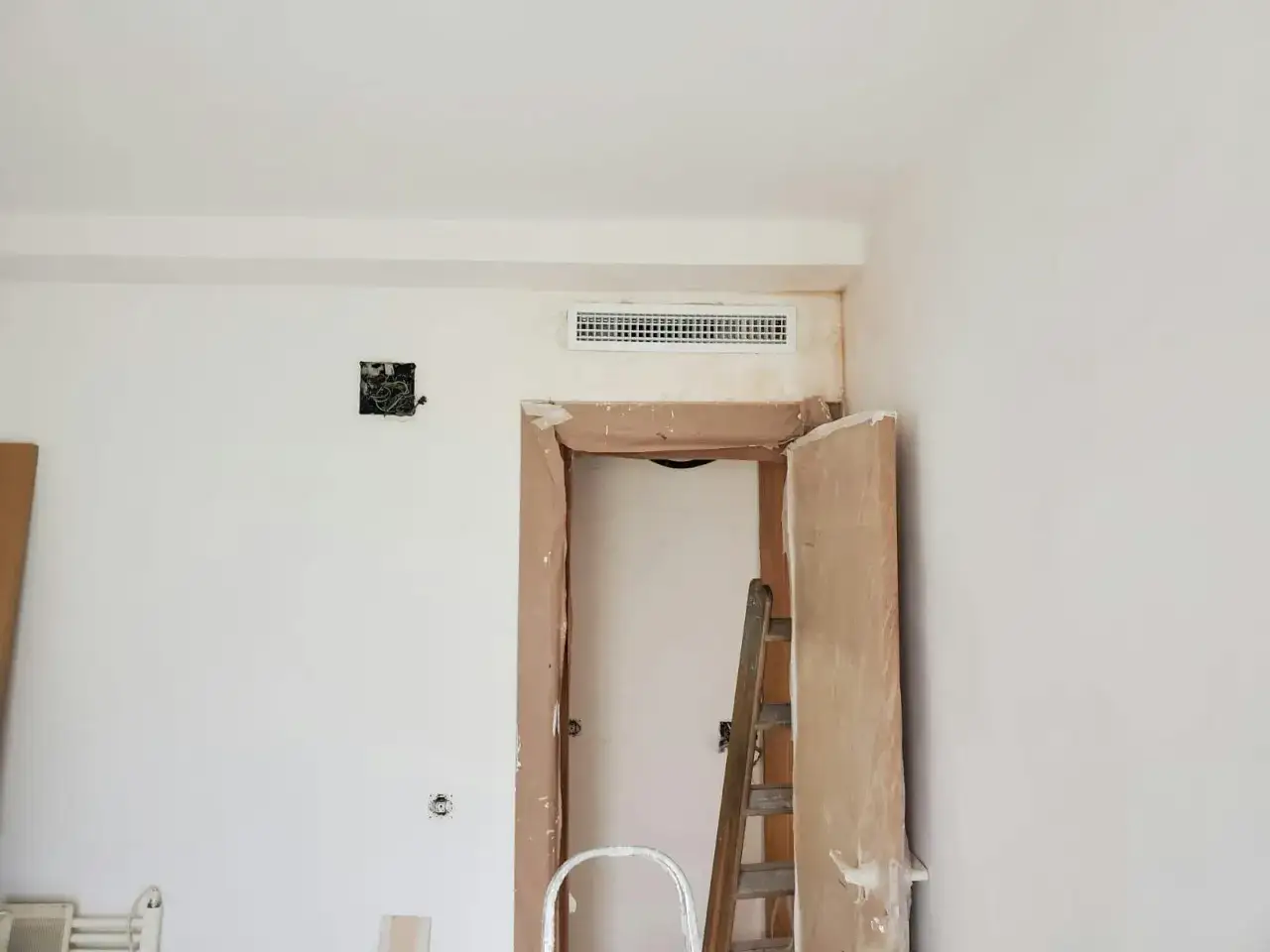 Habitación en obra con preinstalación de aire acondicionado visible en la pared. Una escalera de madera está apoyada en el marco de una puerta.